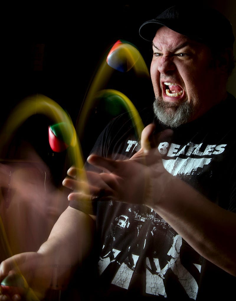 A bearded man juggling hacksacks with an intense, open-mouthed look, suggesting extreme concentration or exasperation